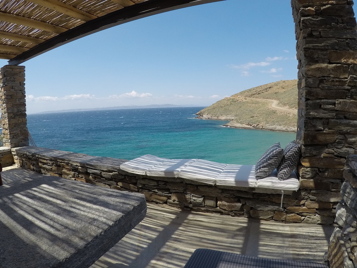 A stone terrace provides a comfortable seating area with large cushions, overlooking the turquoise sea and distant hills. Sunlight creates a striped pattern across the surface, while gentle waves can be seen along the shore, enhancing the tranquil coastal ambiance.