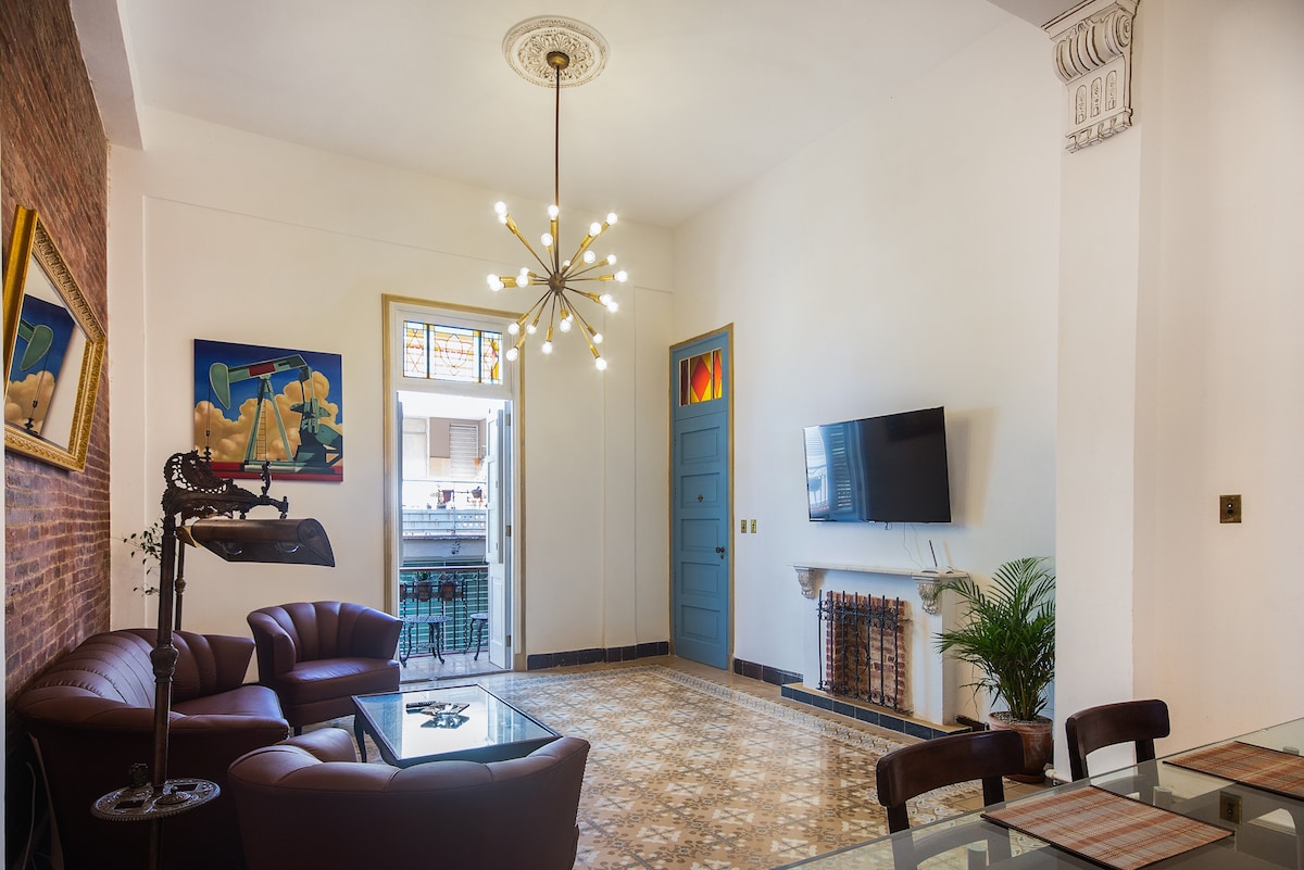 A spacious living area features vintage leather seating and a decorative glass table, enhanced by a modern light fixture. A television is mounted on the wall, and a door opens to an outdoor space. Colorful artwork adds character to the neutral decor.