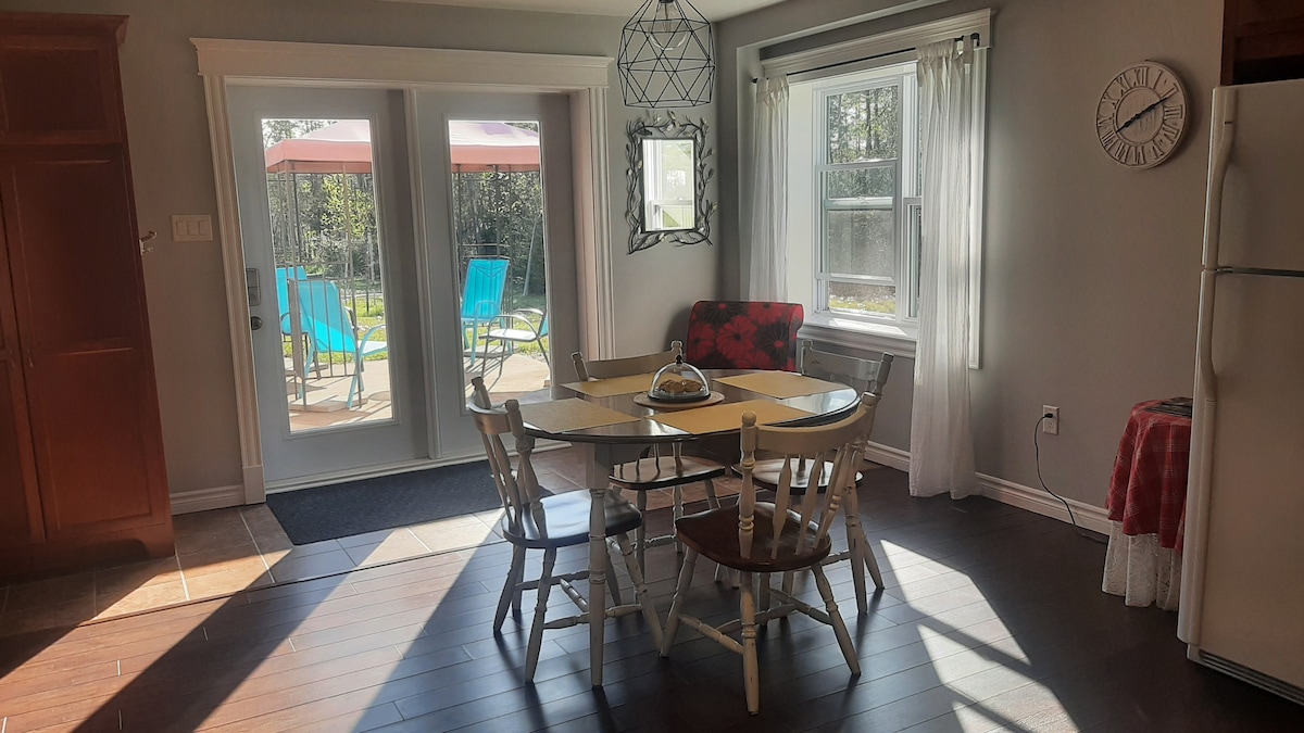 A bright dining area features a round table surrounded by six chairs. Natural light floods the space through glass doors that open to a patio with colorful lounge chairs. A decorative light fixture hangs above the table, while a clock and vibrant seating add character.