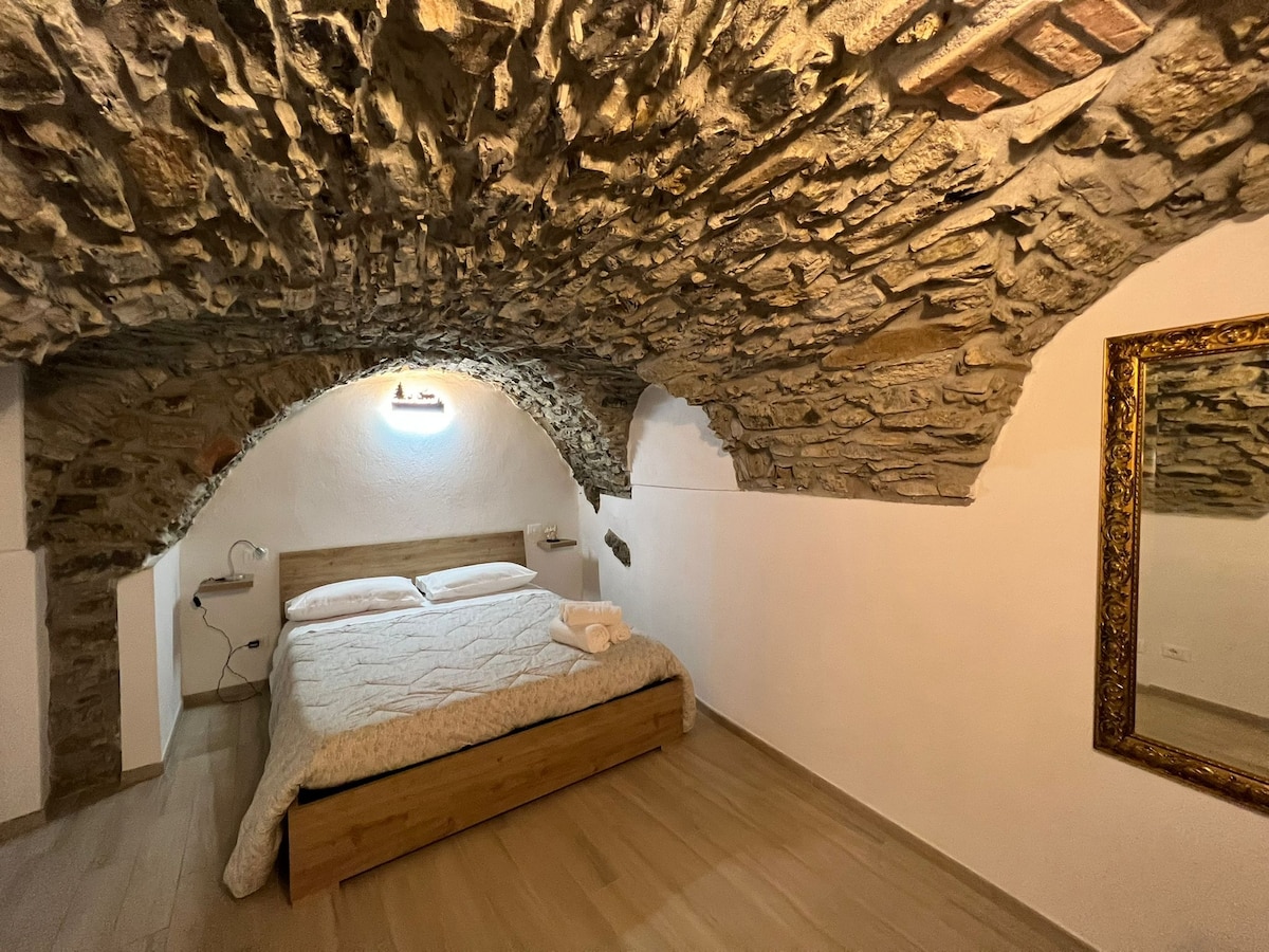 A sleeping area features a spacious bed with neutral bedding set against a backdrop of original stone archways. A decorative mirror reflects the calming ambiance, while soft lighting creates a serene atmosphere within this unique space.