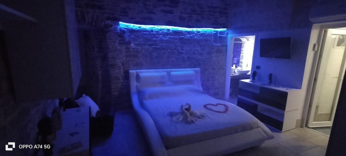 A spacious sleeping area features a modern bed with a soft, rounded design. Soft blue lighting accents the room, highlighting the stone wall. A small entertainment unit and en-suite bathroom are visible, enhancing the contemporary atmosphere.