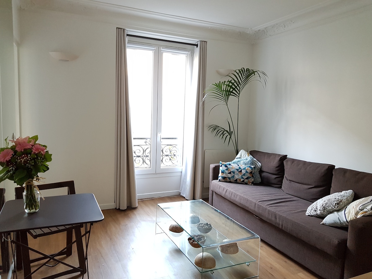 A bright living area features a comfortable brown sofa adorned with decorative pillows. A glass coffee table rests on polished wooden floors, complemented by a small folding dining table nearby. Natural light enters through large windows, framed by curtain panels, with a decorative plant enhancing the space.