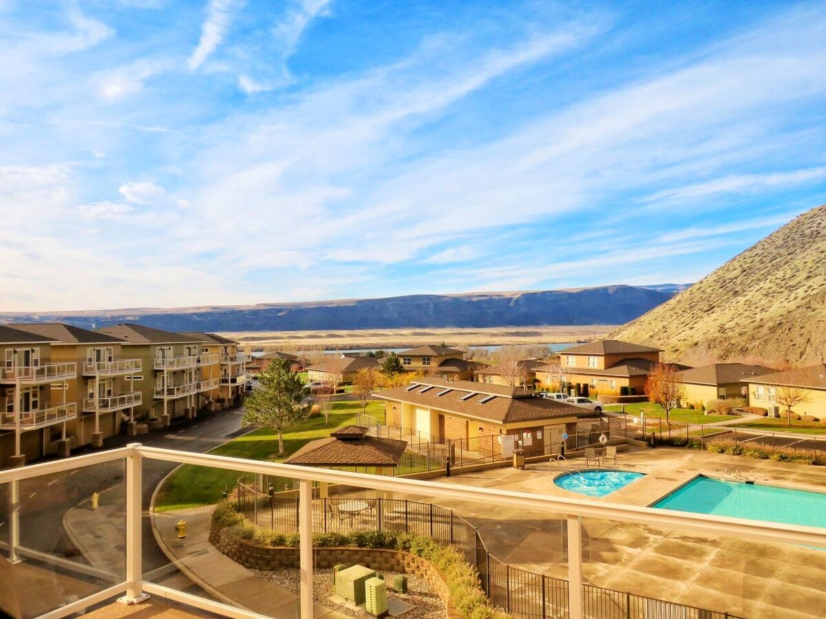 A scenic view from the deck overlooks the expansive canyon landscape, highlighting a swimming pool surrounded by grass and pathways. Nearby, multiple buildings and trees create a calm environment, with distant mountains framed under a bright blue sky adorned with wispy clouds.