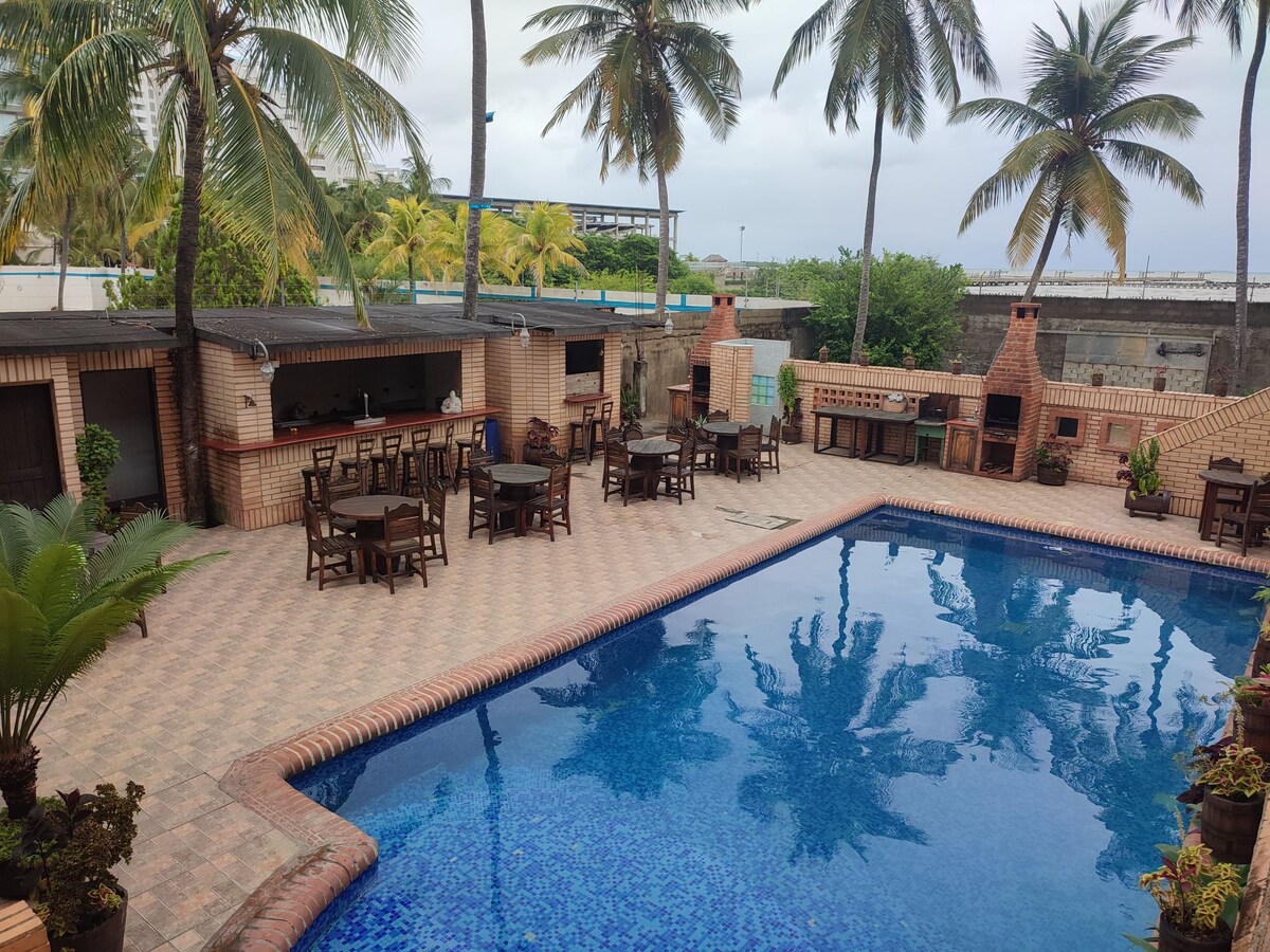 An outdoor pool area is depicted, featuring a spacious swimming pool surrounded by sun loungers and several wooden dining tables. Palm trees provide shade, and a bar area is visible in the background, integrated with a stone structure. The atmosphere appears relaxed and inviting.