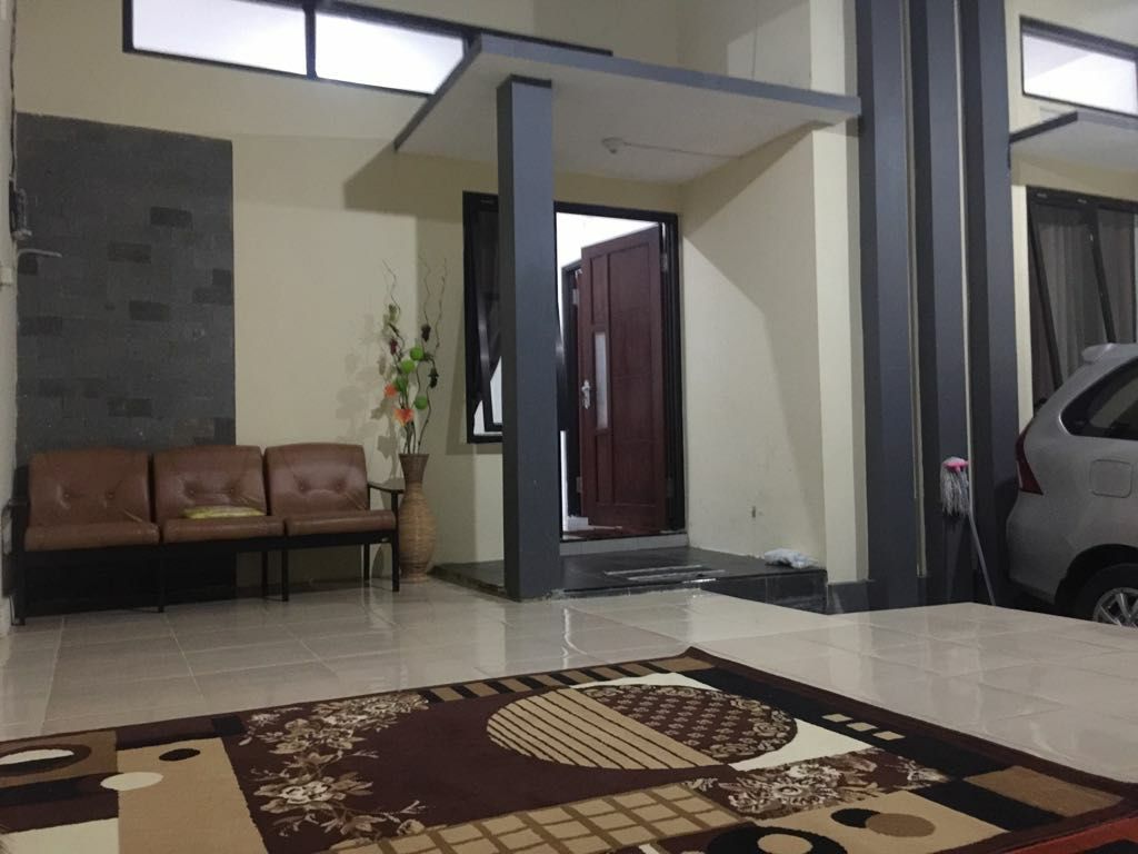 An inviting entrance area features a brown leather sofa paired with a decorative vase. A patterned rug lays on the tiled floor, leading to a door framed by large windows, allowing natural light to fill the space. A car is parked nearby.