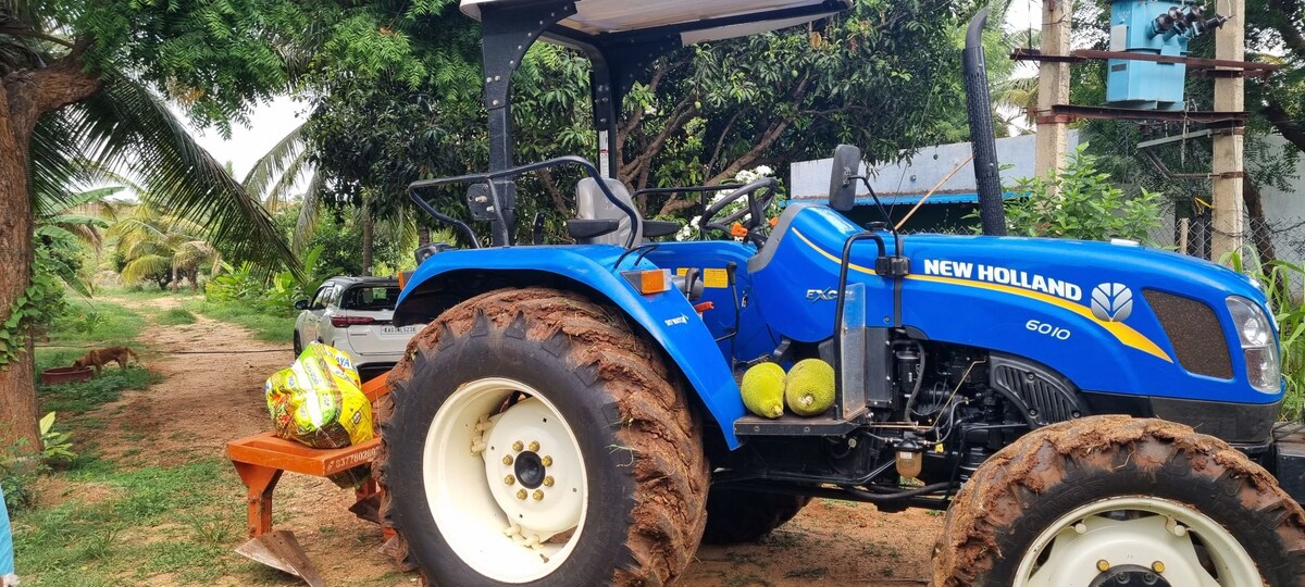 A bright blue tractor is positioned on a dirt path surrounded by greenery. The tractor displays distinct features like large wheels and a roof structure. Nearby, fresh coconuts are visible on a wooden table, contributing to the farm's agricultural atmosphere.