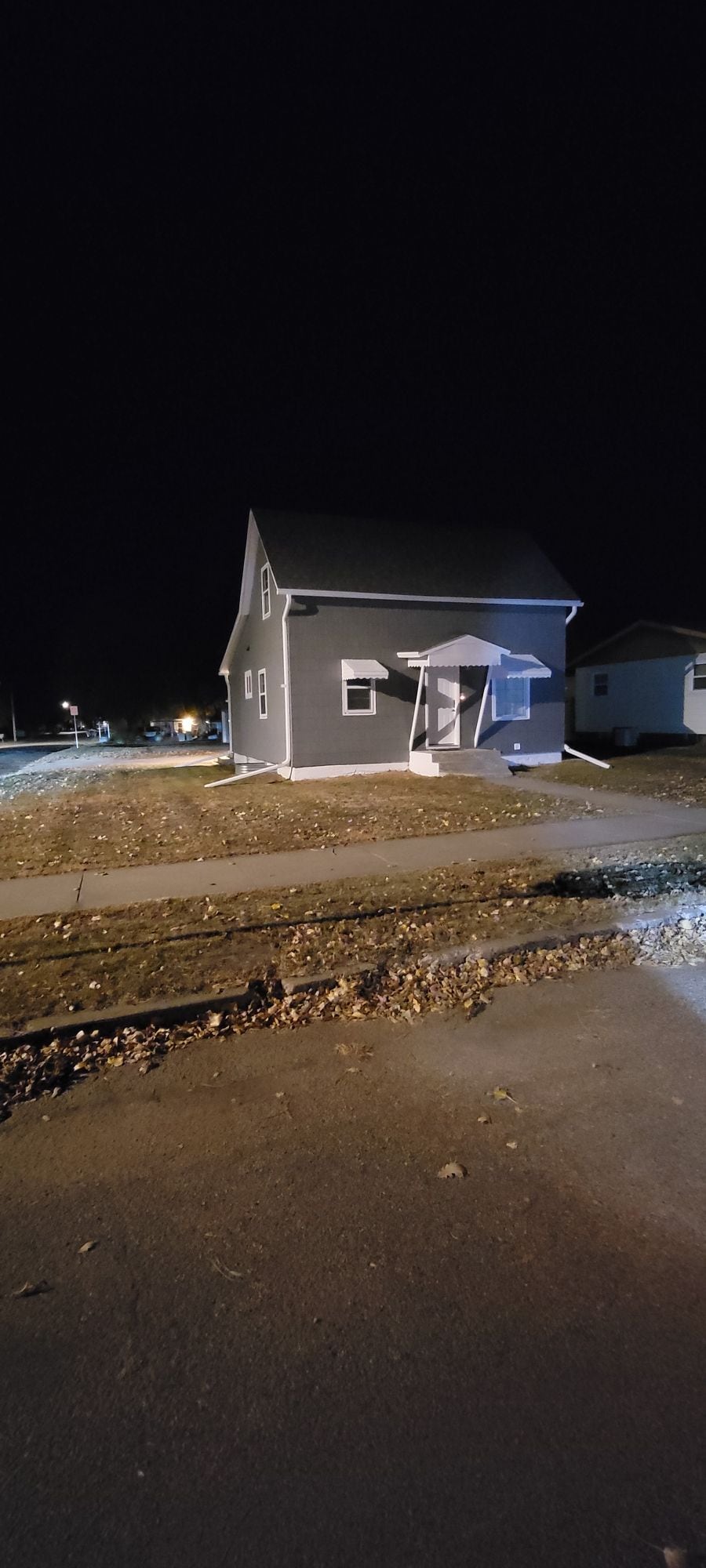 The exterior of a two-story house is shown at night, featuring a light gray facade and a front porch with steps. Soft lighting from nearby street lamps illuminates the surrounding area, providing a sense of safety and visibility.