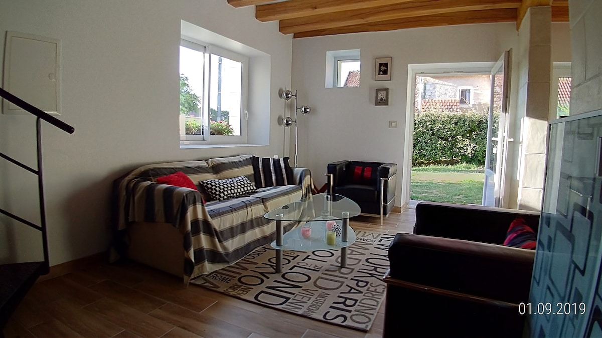 A spacious living area is illuminated by natural light coming through large windows. Two comfortable sofas, one with decorative cushions, are arranged around a glass coffee table. A decorative rug adds texture to the room, while a staircase leads to an upper level.