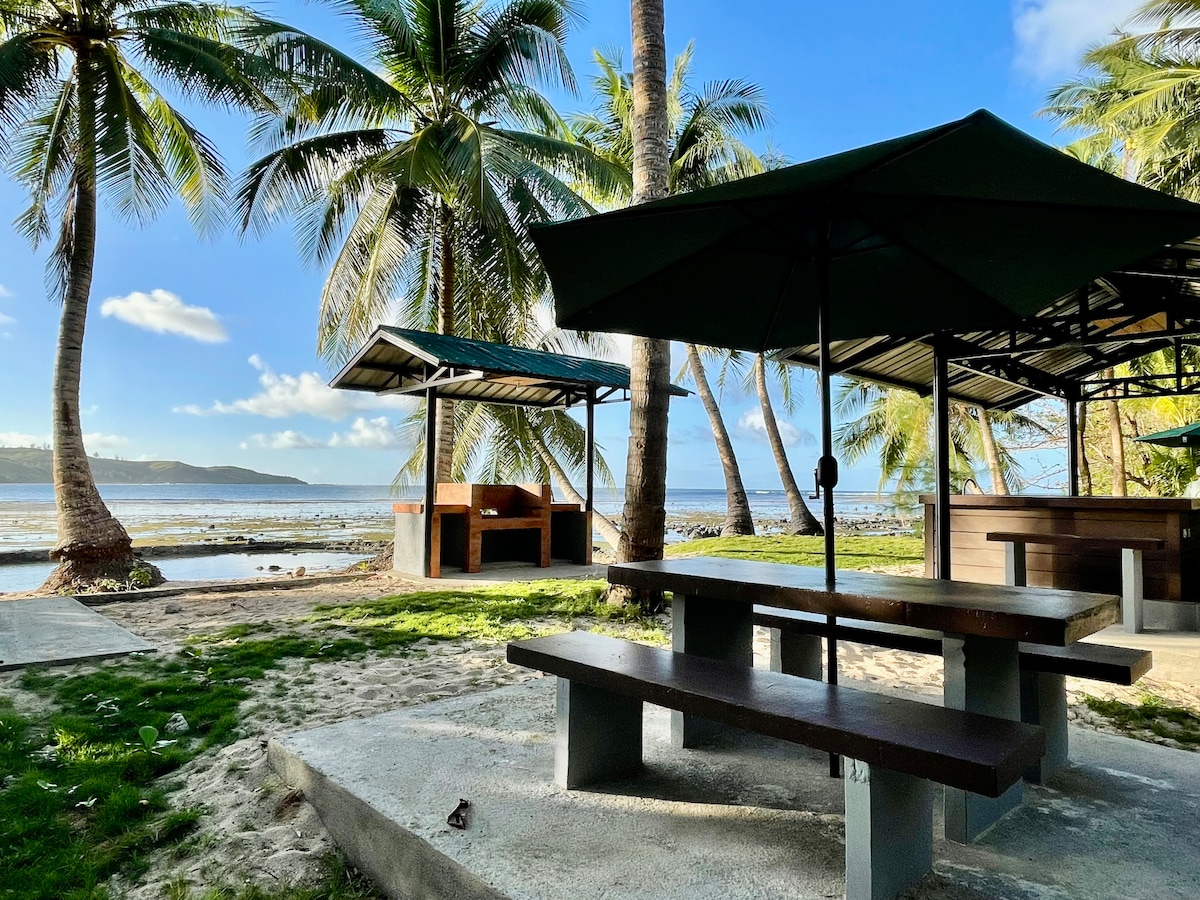 A shaded outdoor dining area provides a table with benches under green umbrellas. Palm trees surround the space, with a sandy path leading to the beach visible in the background. The calm sea and distant shoreline complement the tranquil atmosphere.