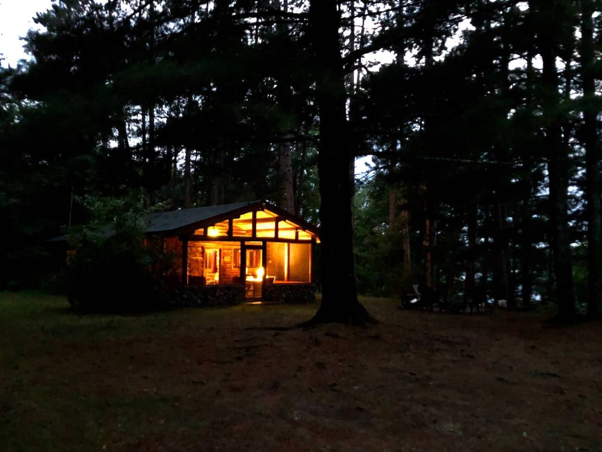 A log cabin is viewed during twilight, illuminated by warm light from the windows. Surrounded by tall trees and greenery, the cabin presents an inviting atmosphere. Natural earth tones of the exterior harmonize with the wooded landscape, offering a sense of solitude and tranquility.