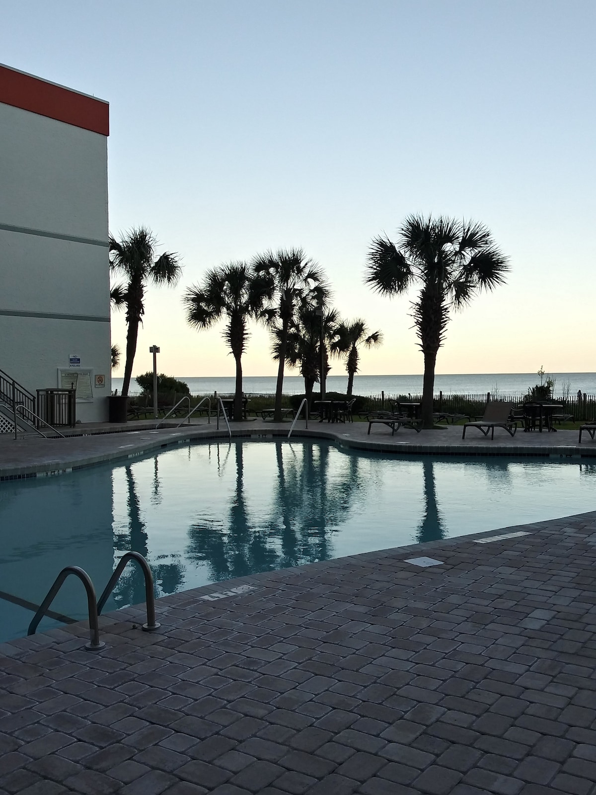 A serene pool area is framed by lush palm trees, with the ocean visible in the distance. The water surface reflects the soft hues of the sky, creating a calming atmosphere. Paved stone surrounds the pool, while lounge chairs are positioned nearby for relaxation.