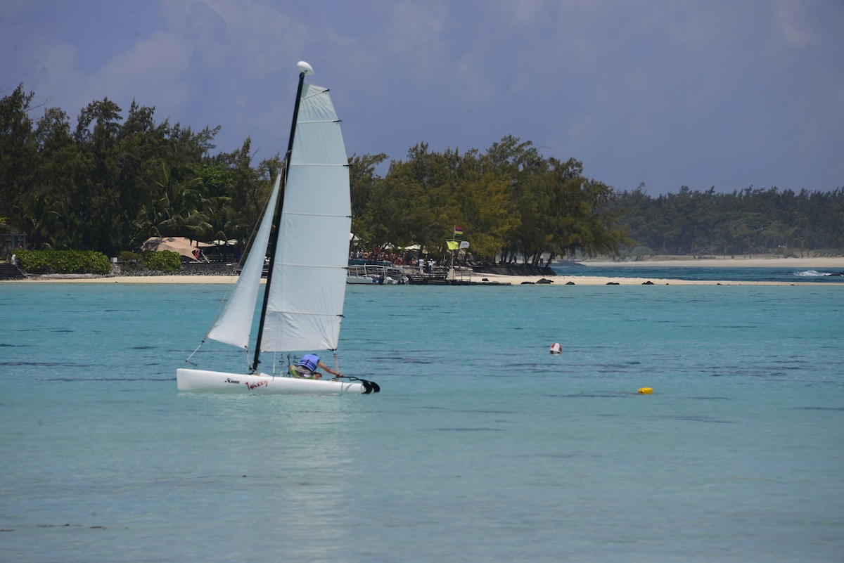 A sailing boat with white sails glides over calm turquoise waters, with gentle waves reflecting the sky. In the background, tropical trees line the shore, and a faint outline of structures can be seen on the coastline, adding to the serene coastal landscape.