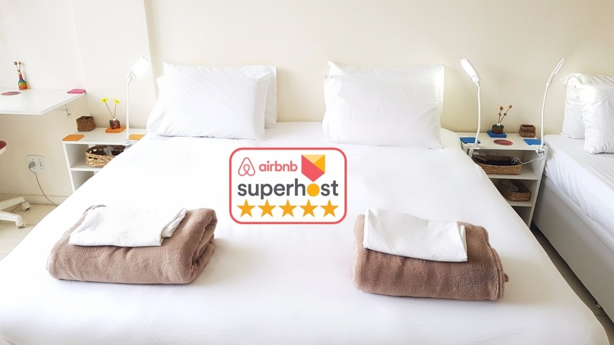 A spacious sleeping area is presented with a large bed dressed in white linens. Soft, folded towels are placed on the bed, alongside bedside lamps and decorative elements on a nearby table. Natural light enhances the clean and minimalist aesthetic.