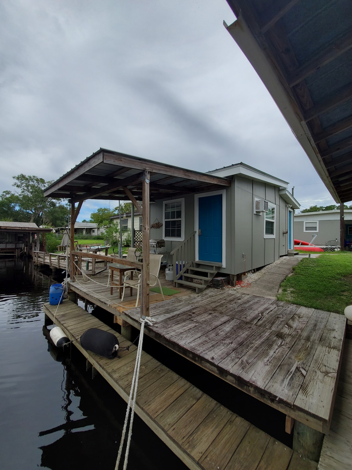 A single-story cottage features a bright blue door and is situated on a wooden dock. The structure is surrounded by water, with a small porch and a couple of chairs inviting relaxation. Lush greenery is visible in the background.