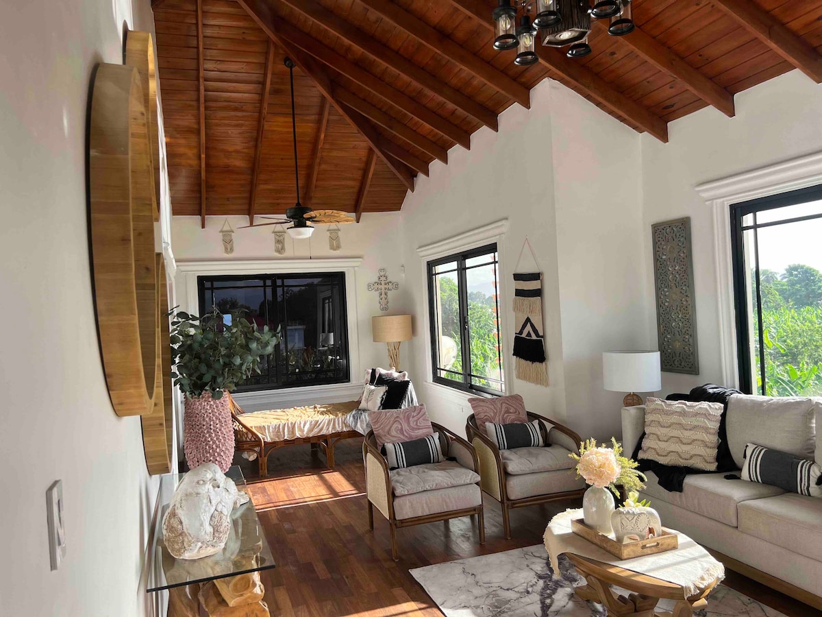 A bright living area features a high, wooden ceiling with thoughtfully arranged furnishings. A comfortable couch is paired with armchairs, and decorative pillows add a touch of comfort. Natural light pours in through large windows, highlighting a coffee table adorned with a floral arrangement.