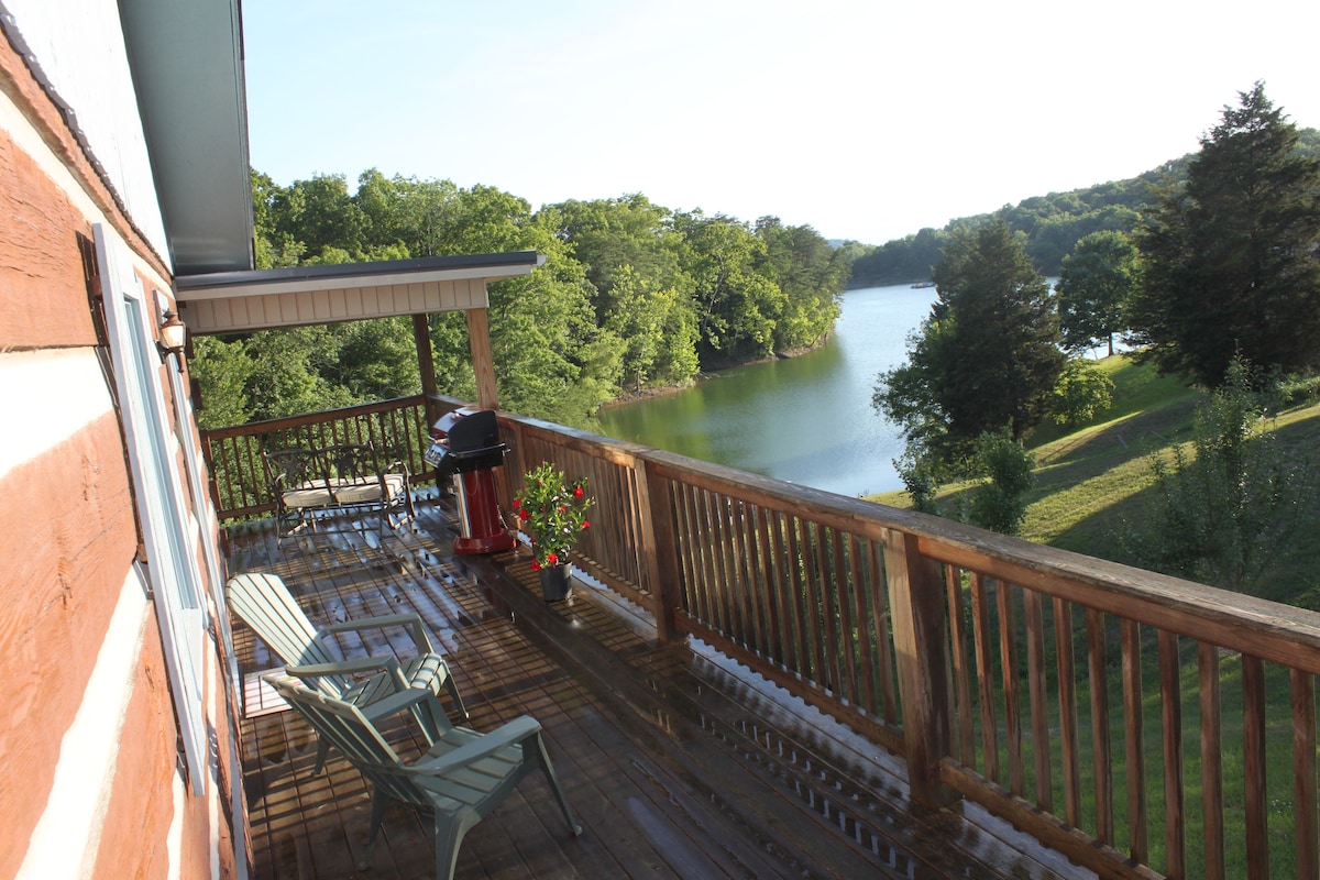 A spacious deck is visible, featuring two comfortable chairs and a gas grill. Lush greenery surrounds the tranquil lake, which reflects the clear sky. The wooden railing offers a scenic view of the water and hillside, creating a peaceful outdoor setting.