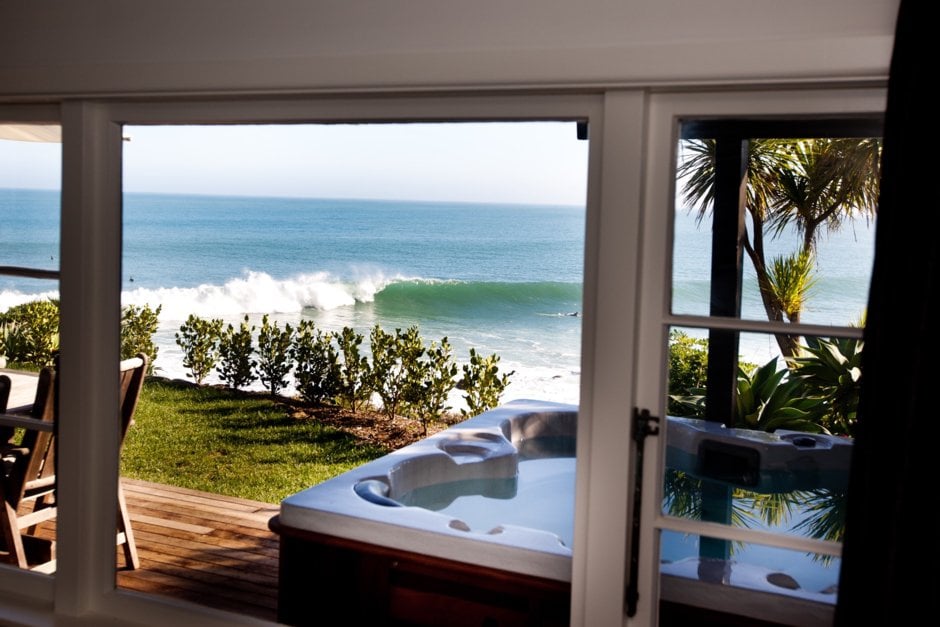 A view from inside showcases a spa pool on a wooden deck, surrounded by greenery. The ocean glistens in the sunlight, displaying gentle waves rolling toward shore. Palm trees frame the scene, highlighting the coastal environment just outside the window.