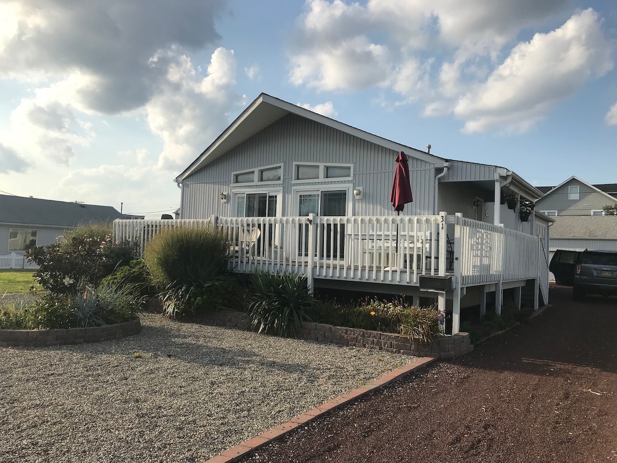 The exterior of a newly renovated beach house is presented, featuring a large front deck with a seating area and a red umbrella. Lush greenery and low-maintenance landscaping frame the entrance, and the structure is characterized by large windows under a gabled roof.