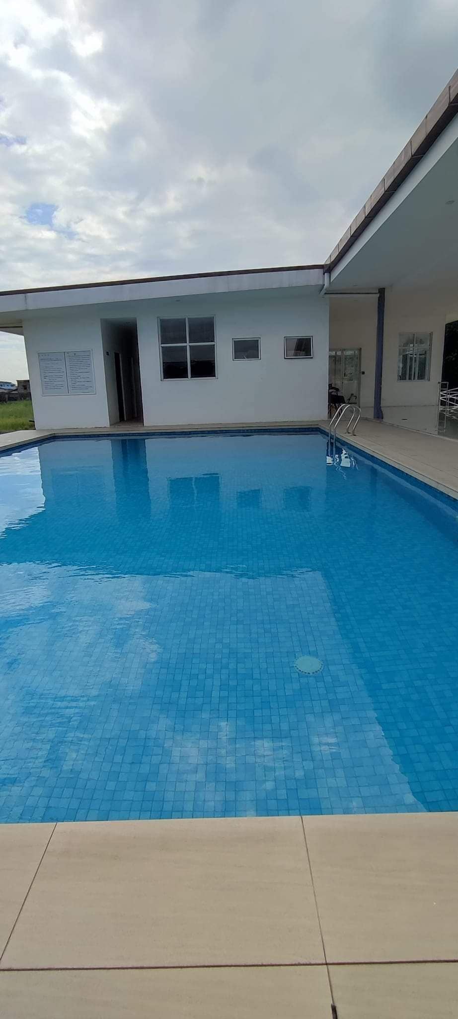 A clear and inviting swimming pool is depicted, featuring bright blue tiles that reflect the sky. Surrounding spaces include a modern building with large windows and a nearby seating area, providing an open and airy environment.
