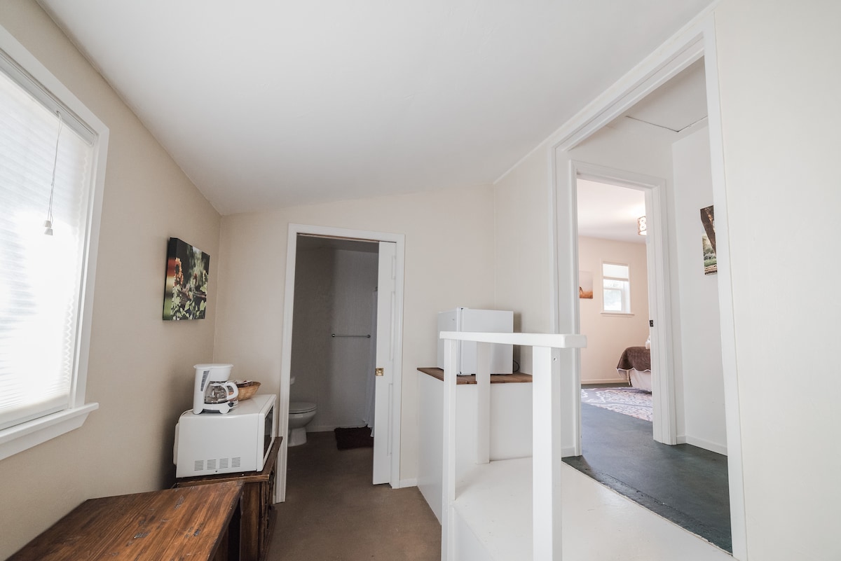 The interior of the space features a light-colored wall and a ceiling with a gentle slope. A wooden side table is positioned against one wall, while a doorway leads to a shared private bathroom. Natural light filters through windows, brightening the area.