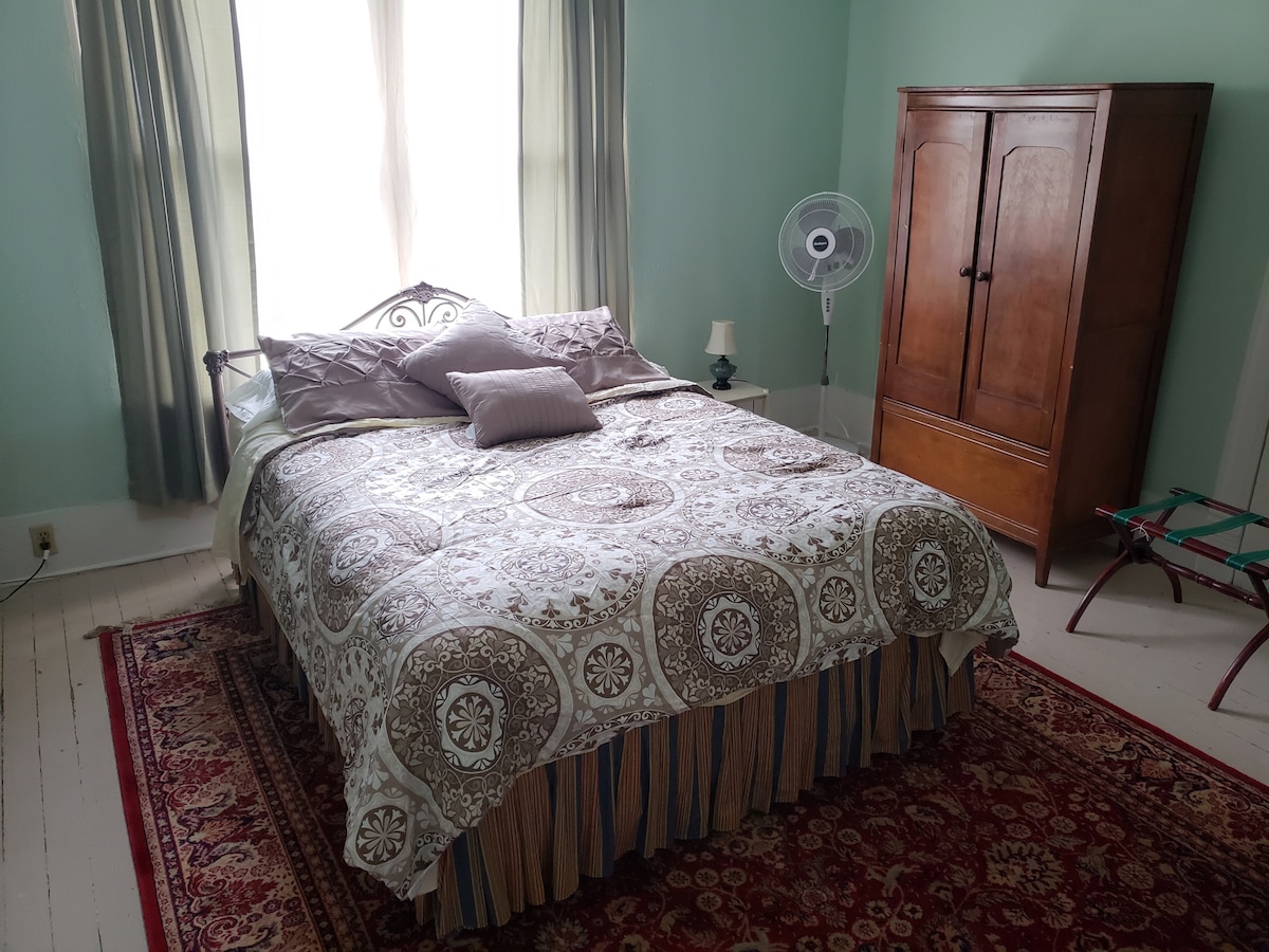 A comfortable sleeping area features a decorated bed with muted tones and several pillows. Natural light enters through large windows, illuminating the space. An armoire stands against the wall, complemented by a decorative rug beneath the bed.