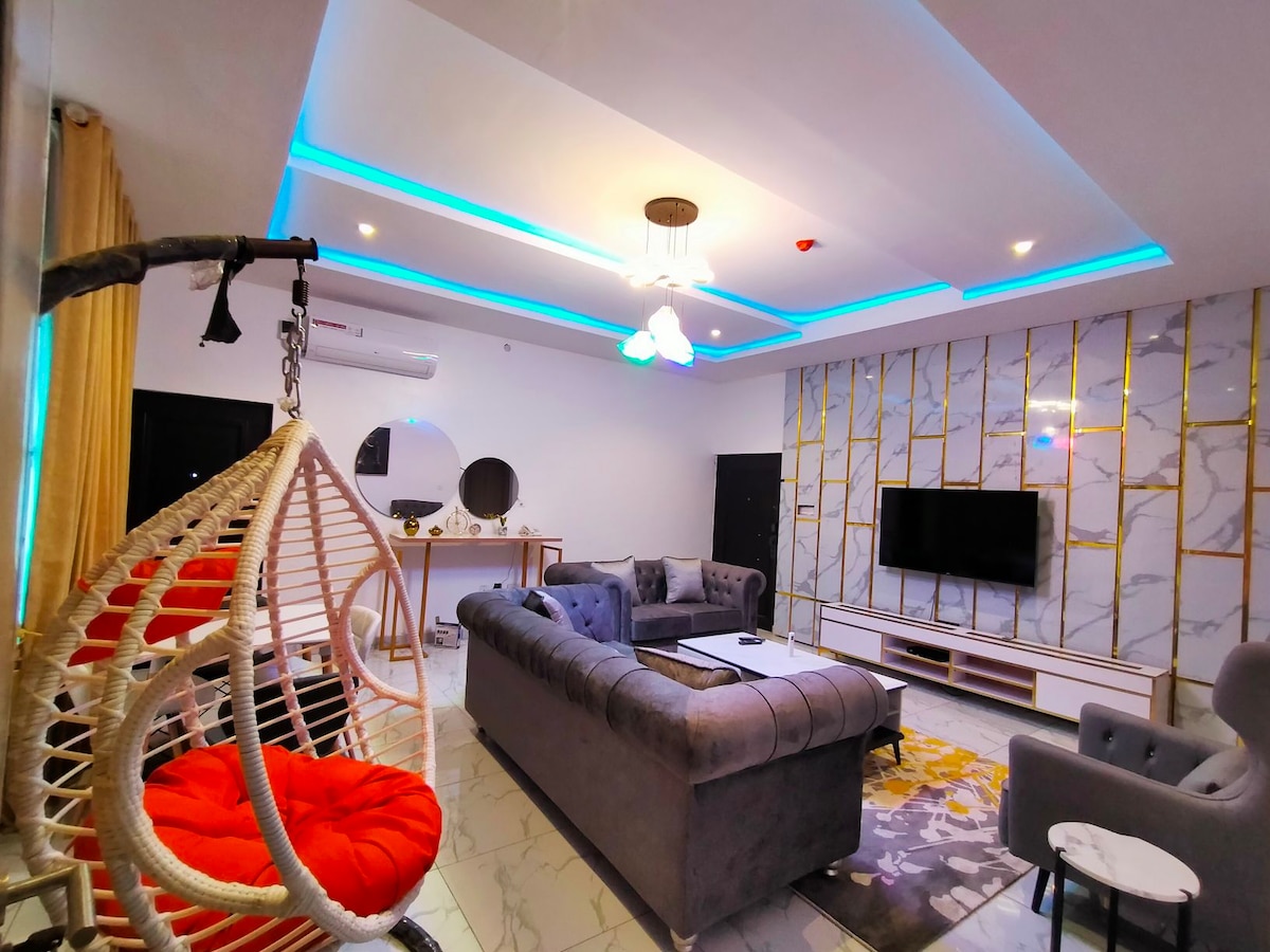 A modern living area is presented, featuring a plush sectional sofa and a stylish hanging chair with a bright cushion. A sleek entertainment unit supports a wall-mounted television. Soft ambient lighting accents the contemporary decor, enhanced by a textured wall and elegant ceiling design.