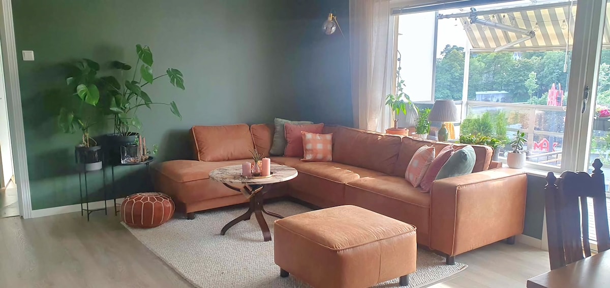 A living area features a large sectional sofa adorned with patterned throw pillows. A round coffee table is centered on a soft area rug. Natural light streams through expansive windows, which overlook a patio area with greenery and outdoor seating.