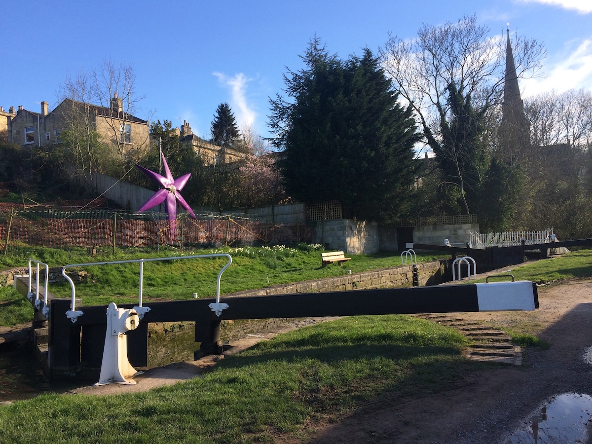 A scenic view of a lock mechanism beside the Kennet and Avon canal, featuring manicured grass and a vibrant pink sculpture in the background. Various trees and buildings provide a natural and urban backdrop, with a church steeple visible in the distance against a clear blue sky.