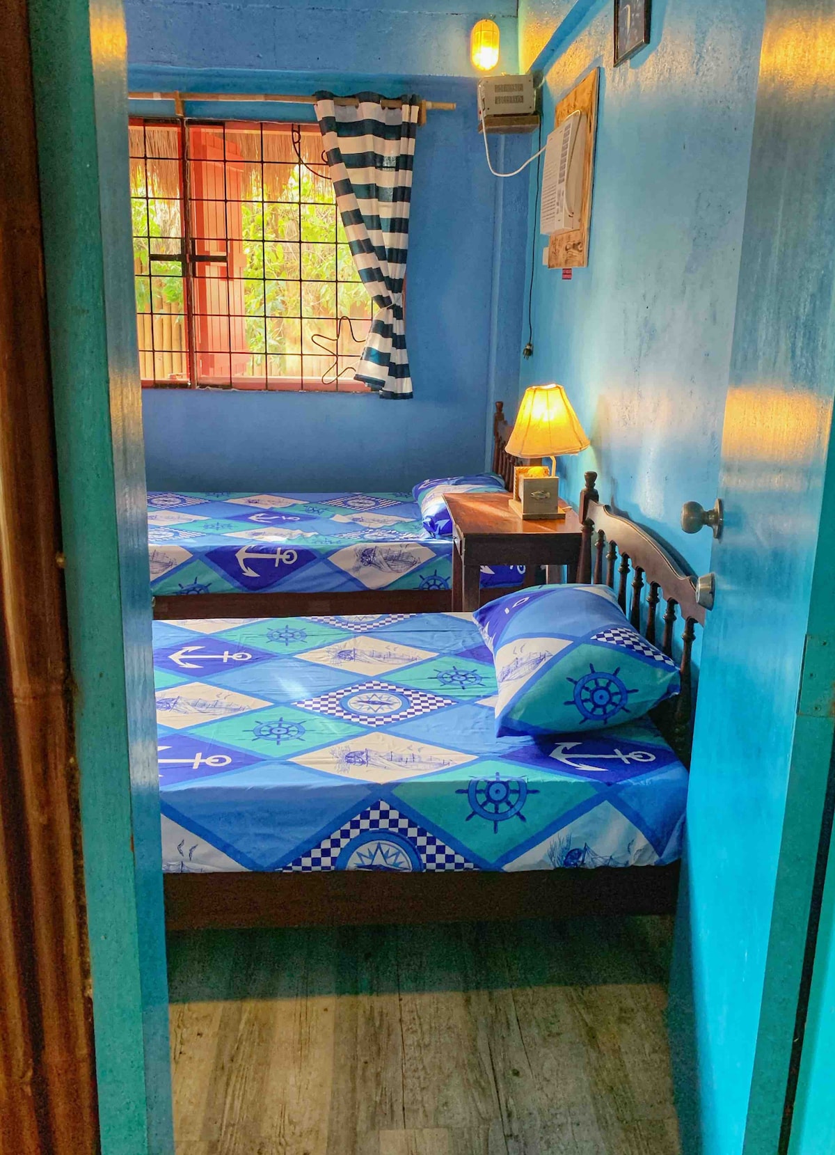 A bright, blue-painted sleeping area features two beds dressed in nautical-themed bedding. Natural light filters through a partially open window with striped curtains, and a bedside lamp adds a cozy touch to the room's decor.