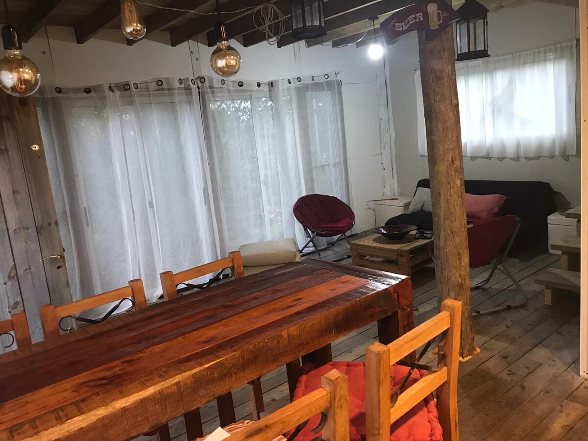 An open living space features a rustic wooden dining table surrounded by chairs. Large windows with sheer curtains allow natural light to fill the room, complementing the warm tones of the wooden flooring and furnishings. A cozy seating area is visible in the background.