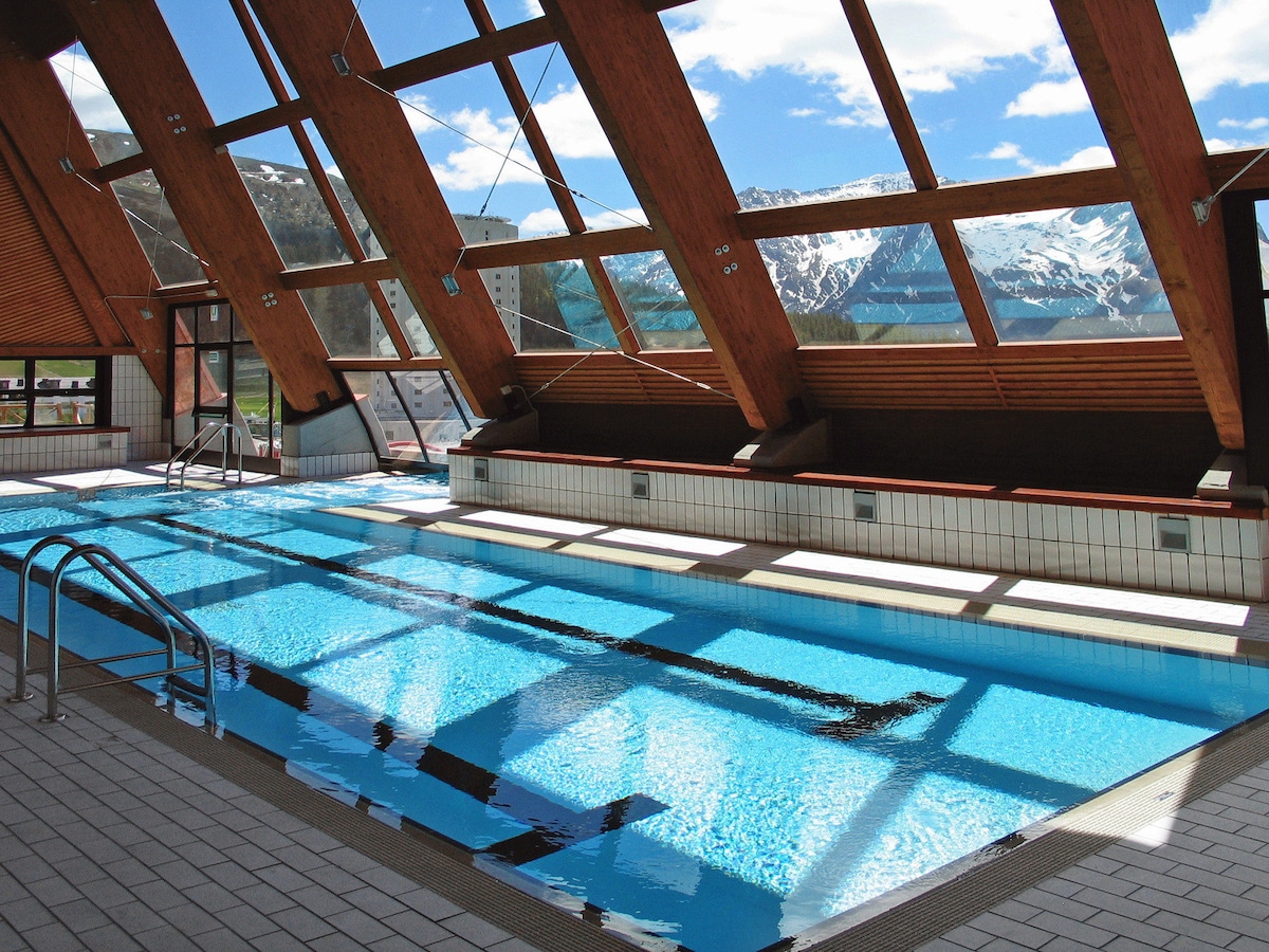 An indoor swimming pool is framed by large windows, allowing natural light to brighten the space. The pool is surrounded by a tiled floor and features modern handrails. Scenic mountain views are visible through the expansive glass installations, creating a serene atmosphere.