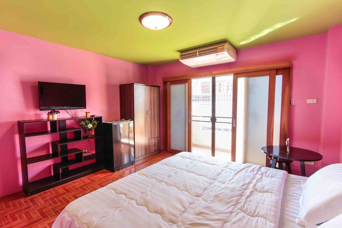 A bright bedroom features pink walls and a vibrant green ceiling. A spacious bed is positioned near sliding glass doors that open to a balcony. A dresser and a mini-fridge are included, along with a small table and a wall-mounted television.