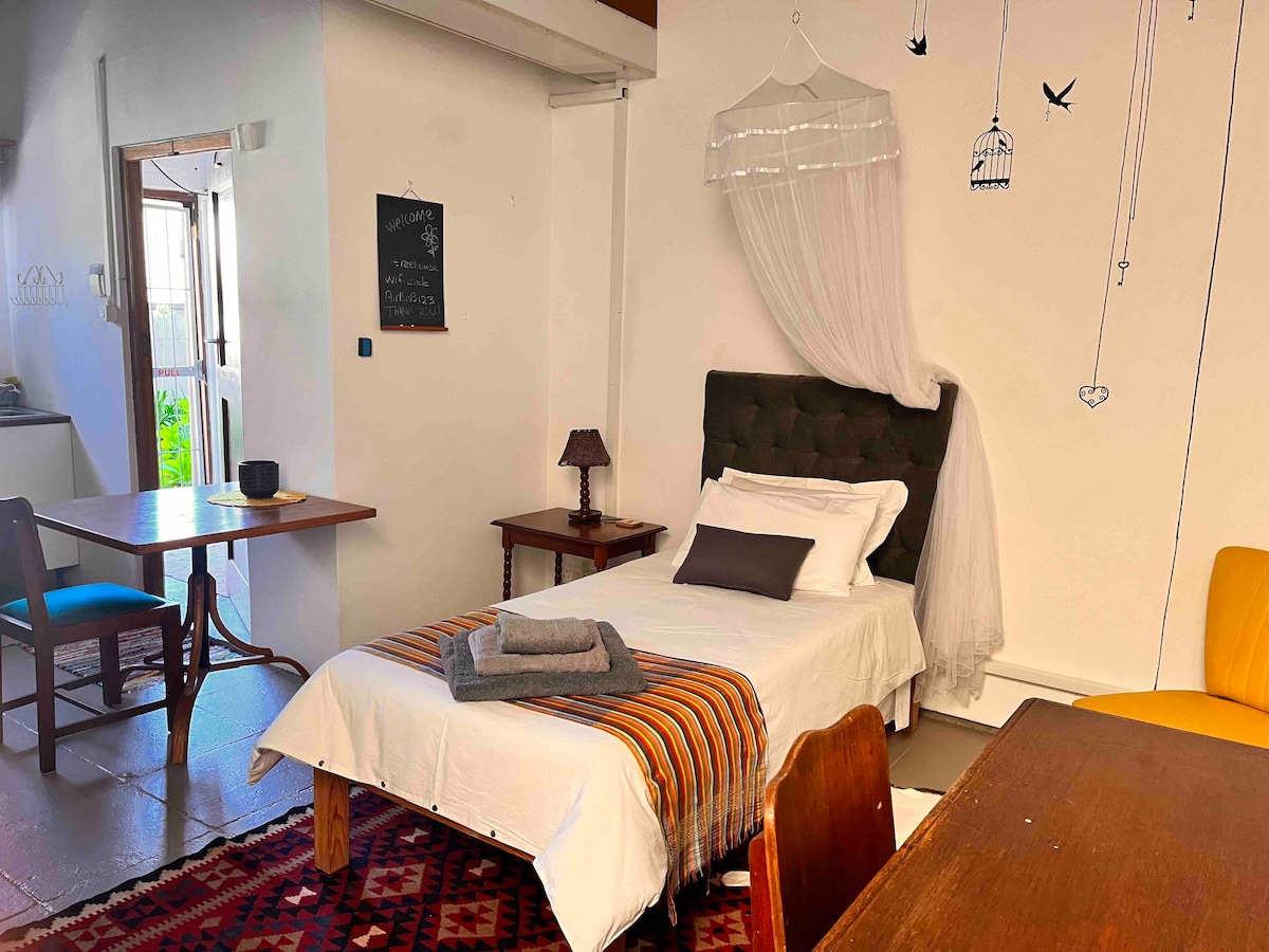 A spacious room is furnished with a neatly made bed featuring a light blanket. A small lamp sits on a wooden table beside the bed. An additional table and chairs occupy the room, with decorative wall art and a light curtain adding soft detail.