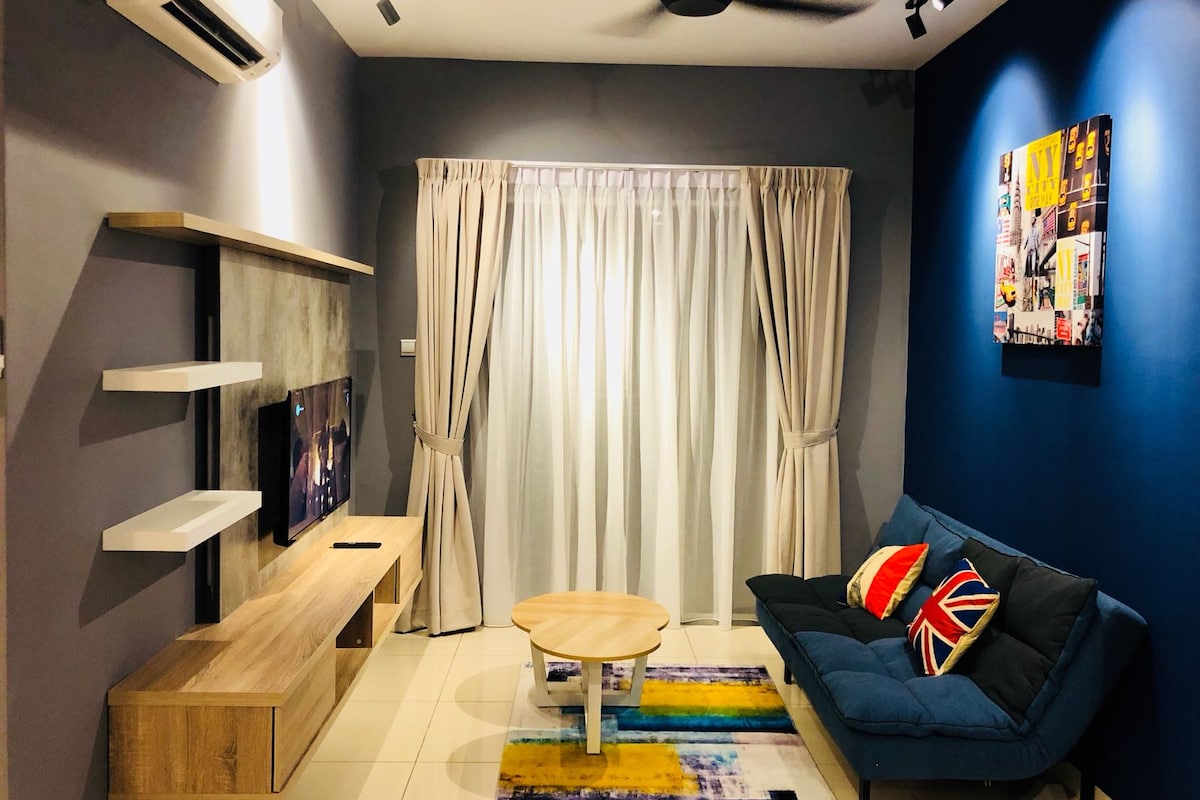 The living area features a comfortable sofa adorned with colorful cushions, complemented by a wooden coffee table. Light-colored curtains frame a window, allowing natural light to fill the space. A television is mounted on a simple, contemporary wall unit, enhancing the inviting atmosphere.