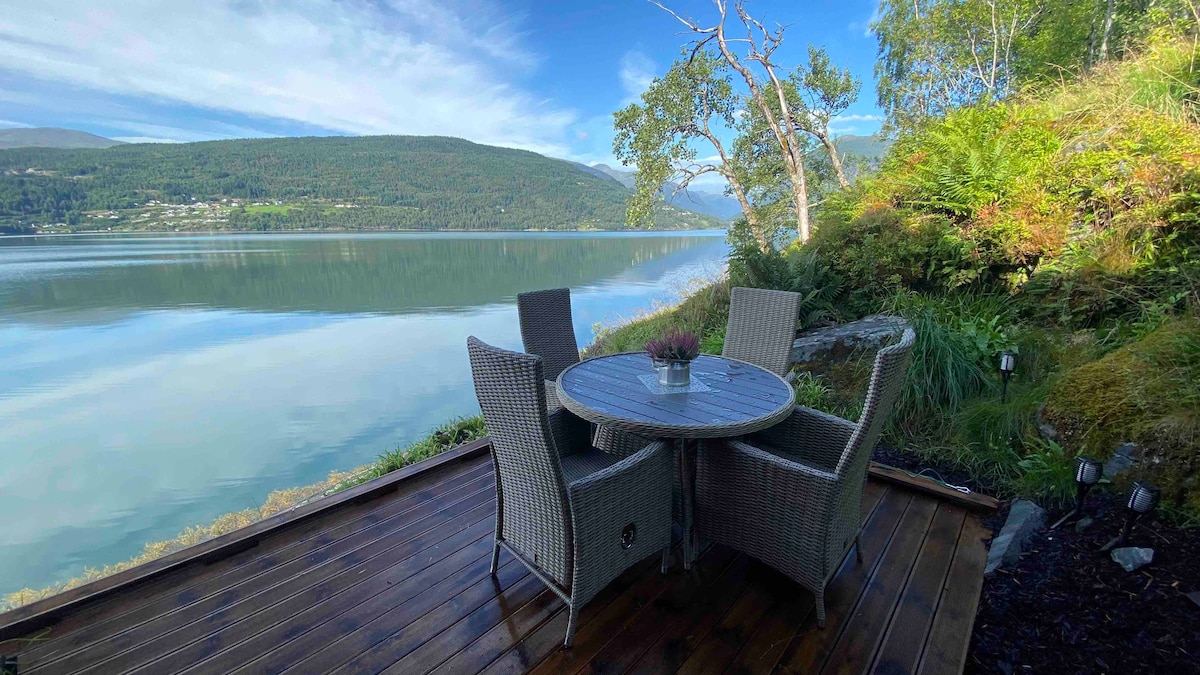 A round table with four wicker chairs is situated on a wooden deck overlooking a serene body of water. Lush greenery and trees are visible in the background, and the calm surface of the water reflects the surrounding landscape.