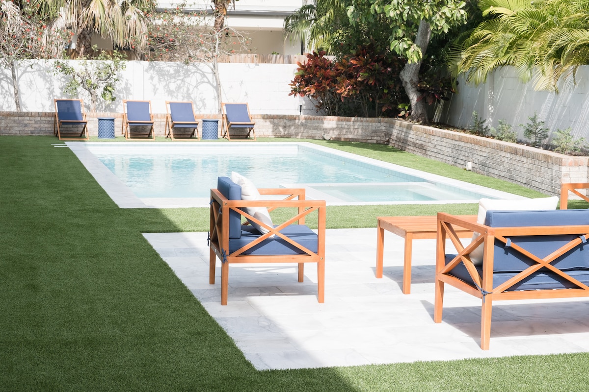 A well-maintained outdoor area features a heated swimming pool surrounded by lush green grass. Lounge chairs with blue cushions are arranged around the pool, alongside wooden tables. Tropical plants provide natural privacy at the edge of the space.