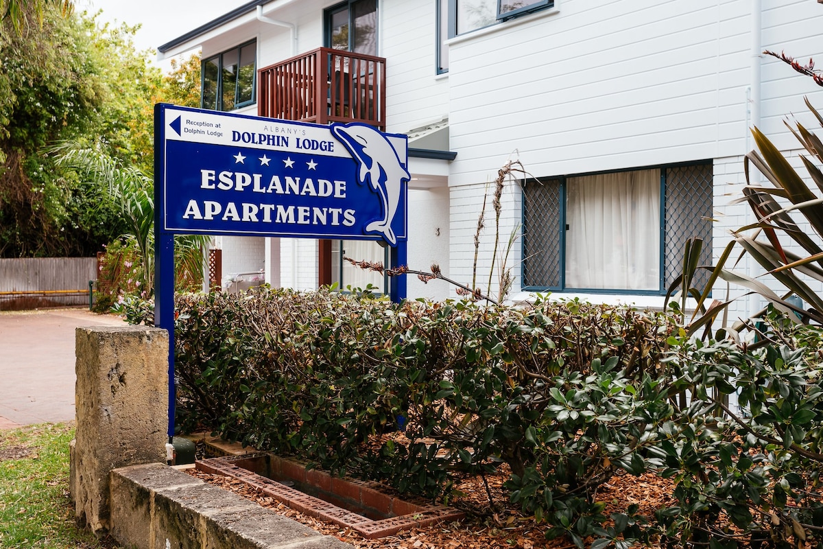 The image showcases the entrance sign of the Esplanade Apartments, featuring an outlined dolphin logo. Surrounding foliage and a landscaped garden area enhance the welcoming appearance of the property, which has a contemporary building structure in the background.