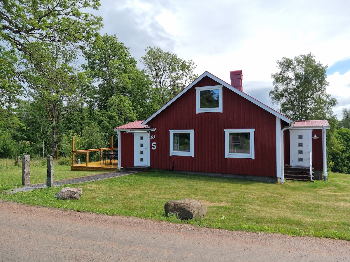 A charming red cottage is set against a backdrop of lush greenery. The building features white accents and multiple windows, enhancing its inviting exterior. A wooden deck extends from the front, leading to a well-maintained lawn and a gravel pathway along the roadside.