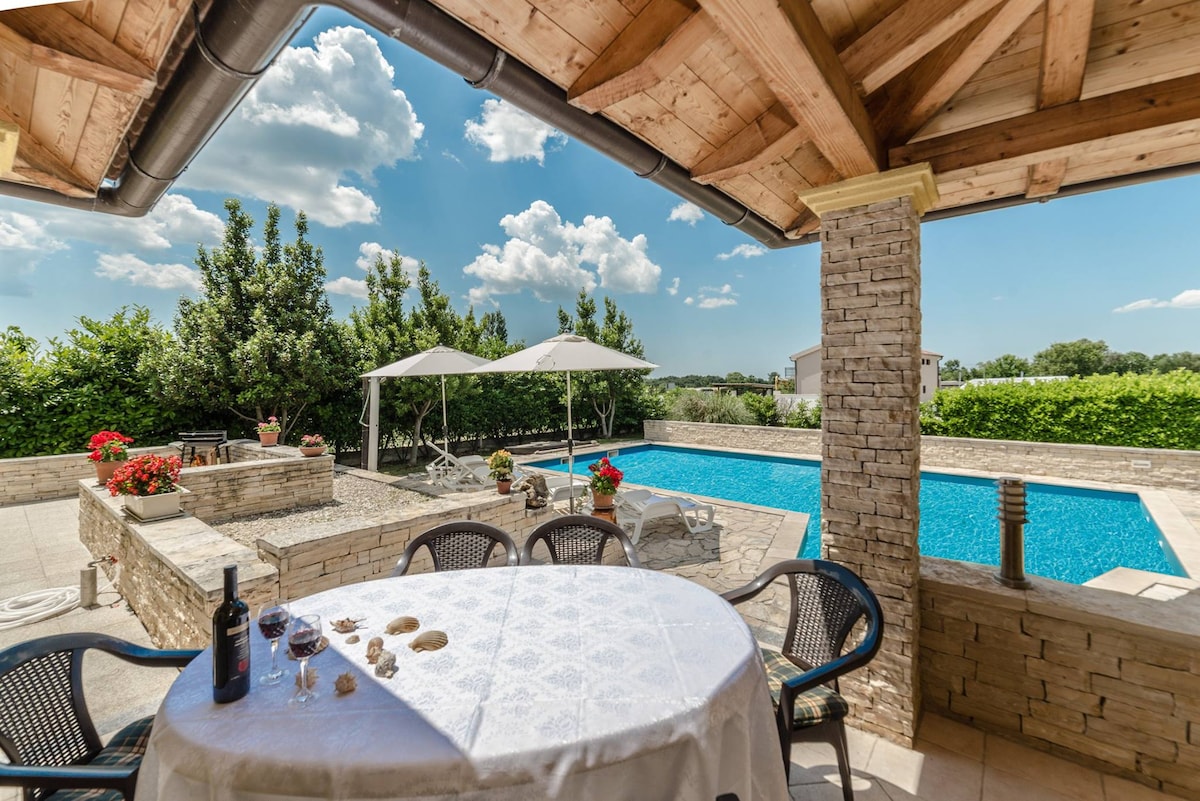 A covered terrace features a round table set with a white tablecloth and two glasses of wine, overlooking a large swimming pool. The pool area is surrounded by landscaped greenery and sun loungers, complemented by shaded seating under white canopies.