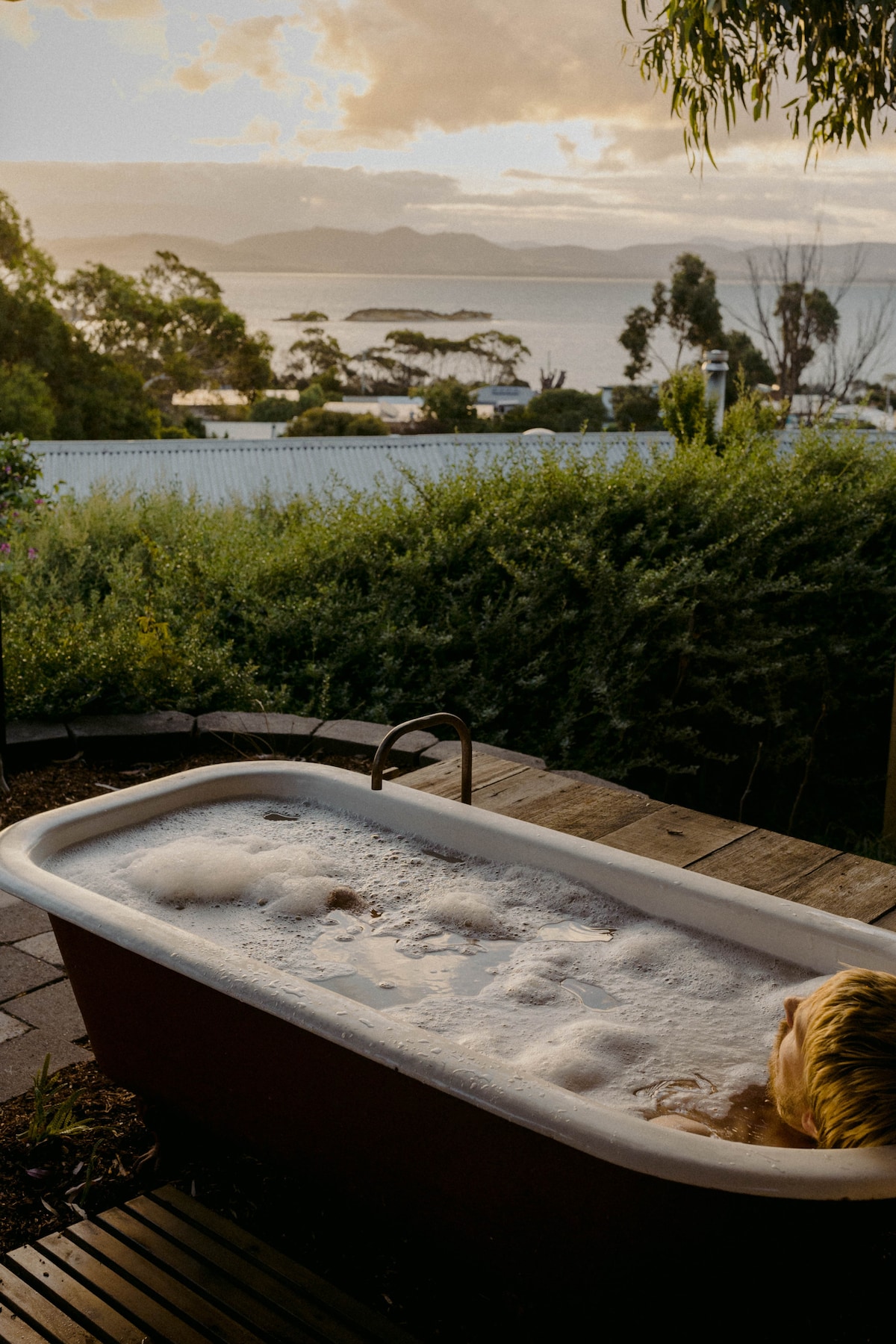 An outdoor bathtub overlooks a serene landscape with distant islands and ocean views. Bubbles are visible in the tub, while lush greenery surrounds the area, enhancing the natural setting. Soft light casts a warm glow over the scene, indicating a peaceful evening.