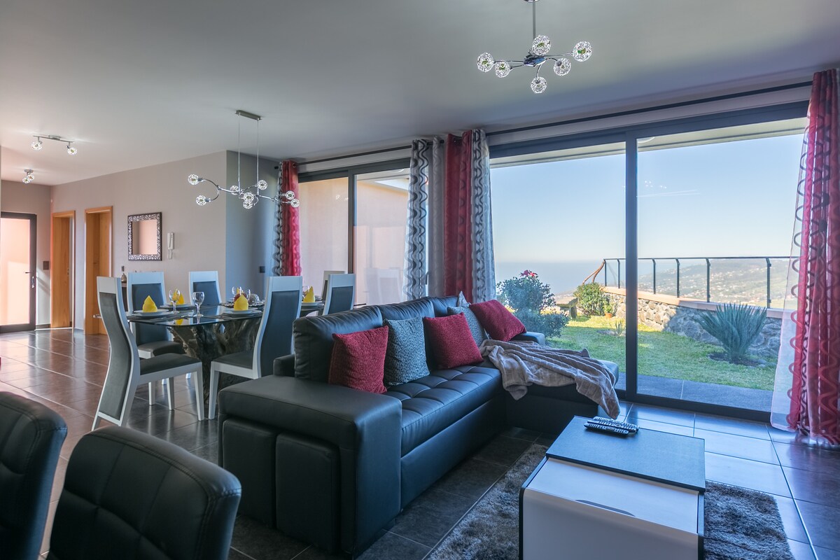 A spacious living area is presented with large glass doors offering unobstructed views of the surrounding landscape. A comfortable black sectional sofa is accompanied by decorative pillows, while a dining table is set for four. Natural light illuminates the space, complemented by modern lighting fixtures.