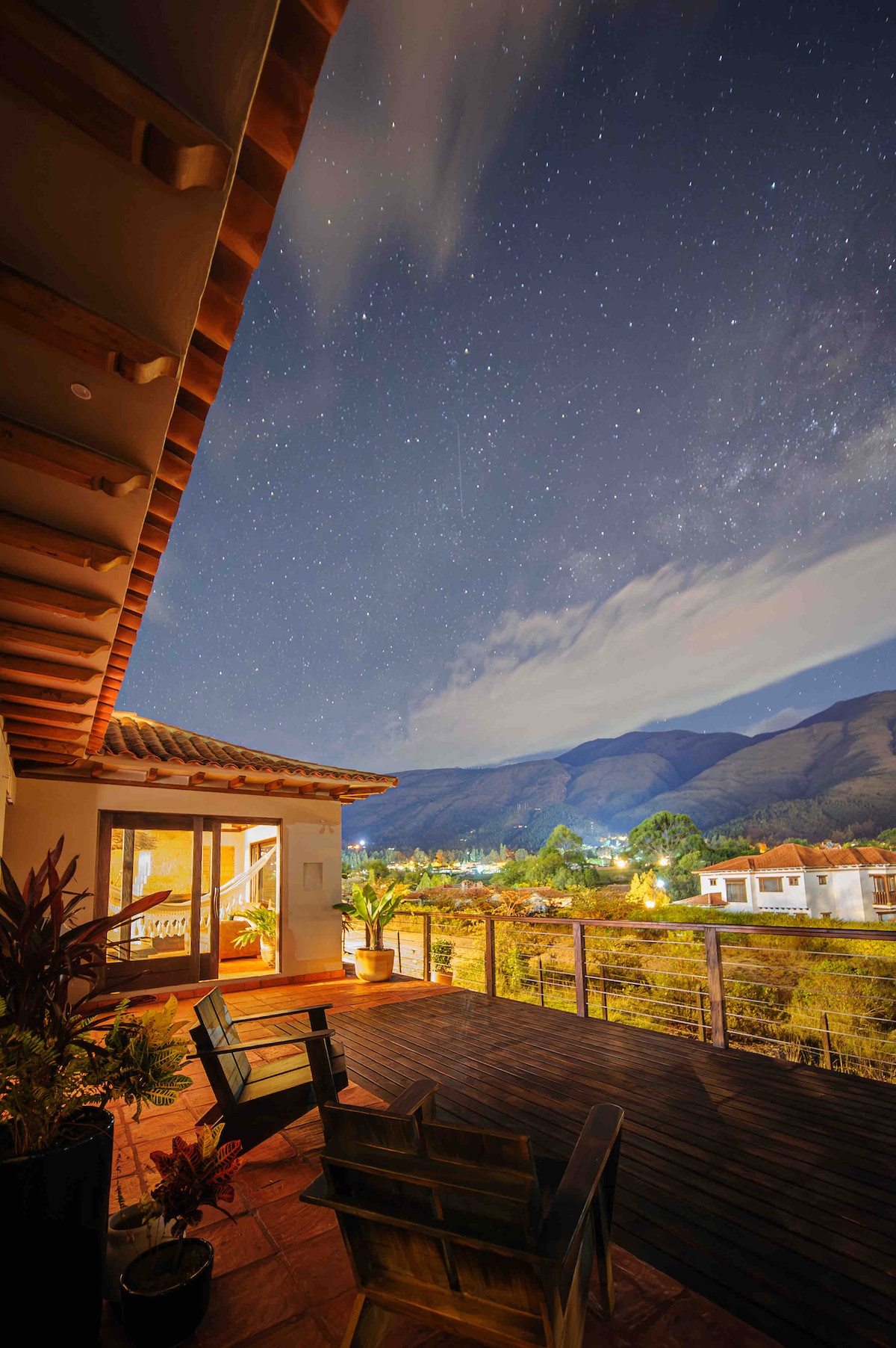 A spacious terrace features two wooden chairs, allowing for a perfect view of the starlit sky and surrounding mountains. Soft lighting from the interior creates a welcoming contrast to the natural scenery, enhancing the tranquil atmosphere.