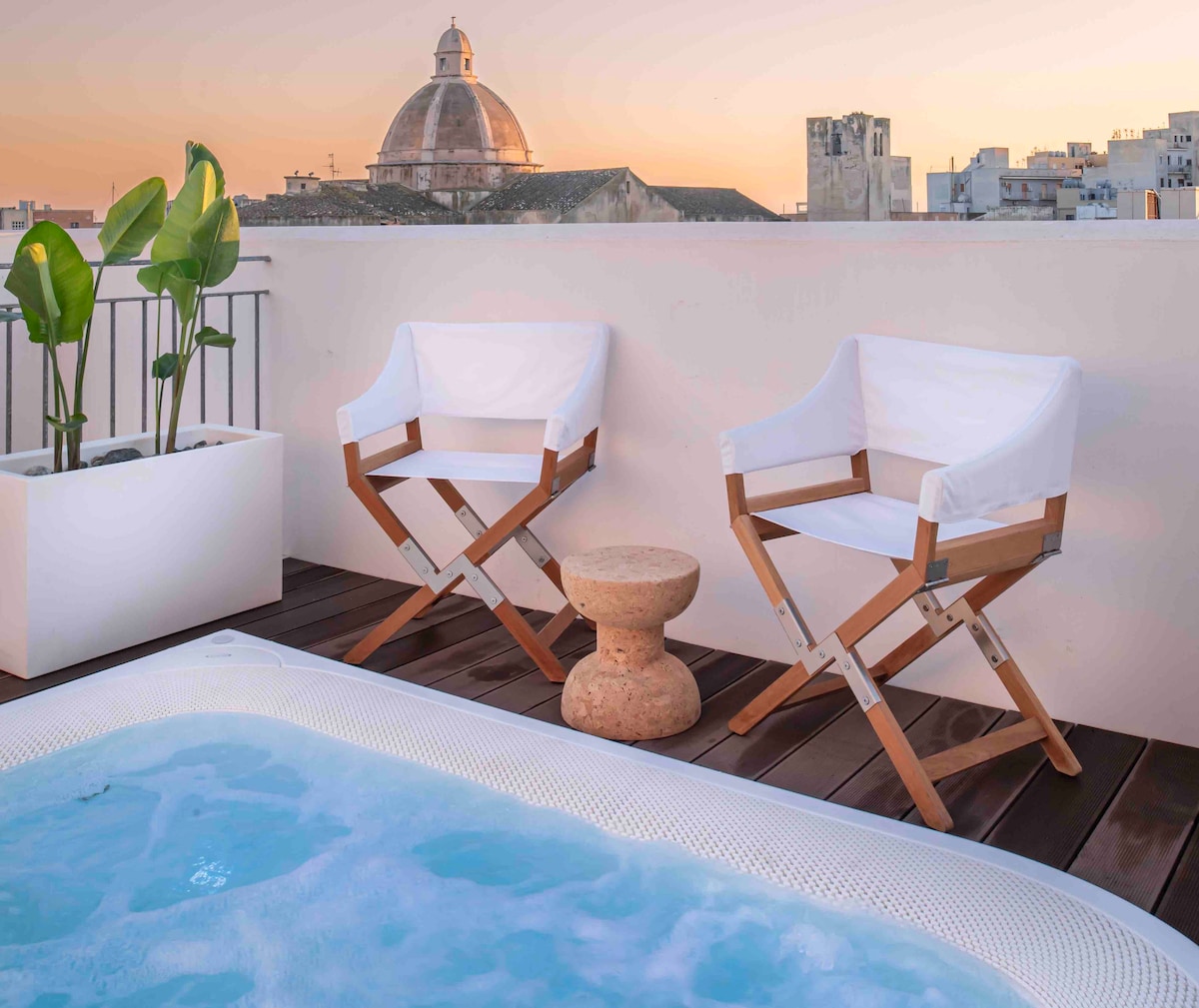 A rooftop terrace is presented with two white folding chairs positioned beside a small stone table. A Jacuzzi is nearby, and the soft glow of sunset highlights the cityscape, with a domed building visible in the background.