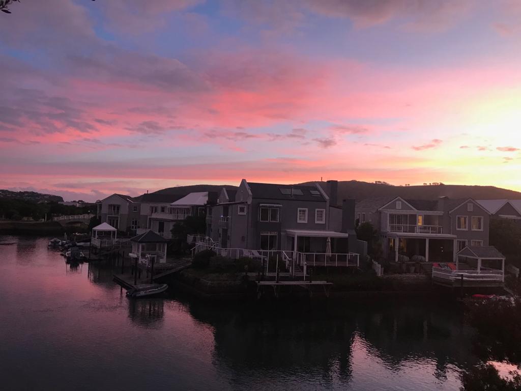A serene view of waterfront homes reflects against the calm water of the canal. The sky displays a gradient of soft pink and purple hues during sunset, enhancing the peaceful atmosphere of the estate.
