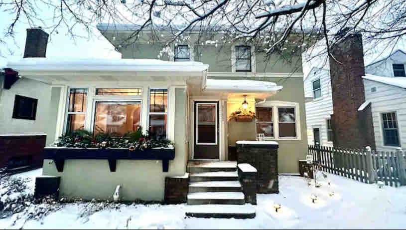 A charming home with distinctive architectural features is framed by a light dusting of snow. The front porch is decorated with greenery, and softly glowing lights illuminate the snowy pathway leading to the entrance. The classic facade highlights original design elements.