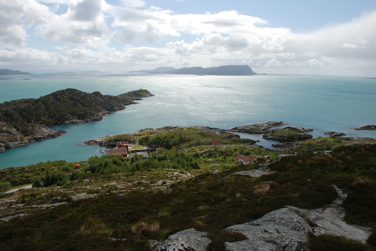 A scenic view of the coastline showcases a variety of rocky islands surrounded by shimmering blue waters. The landscape features patches of greenery, and distant hills can be seen under a partly cloudy sky, creating a tranquil setting.