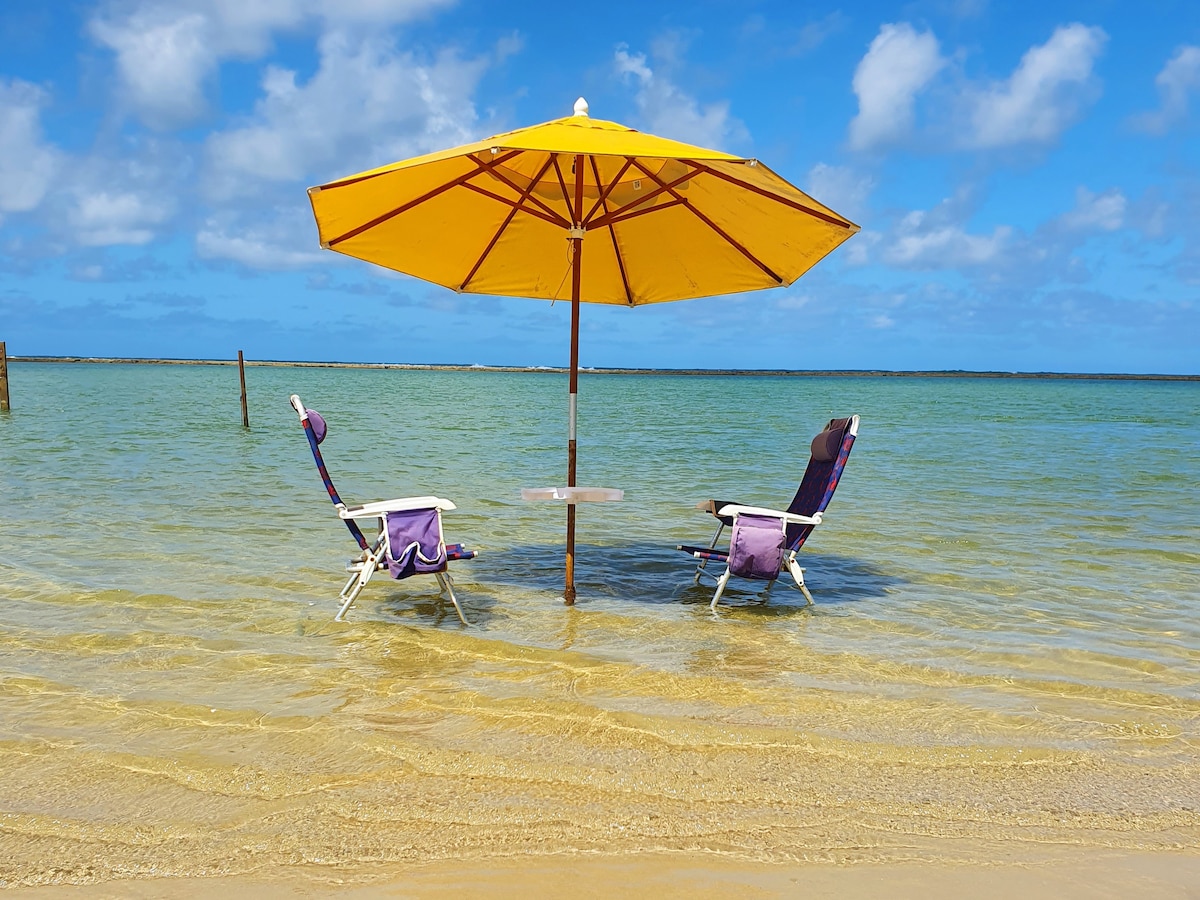 Two beach chairs are positioned beneath a large yellow umbrella, partially submerged in clear, shallow water. A small table is set between the chairs, all overlooking the tranquil sea, with blue skies and a few fluffy clouds in the background.