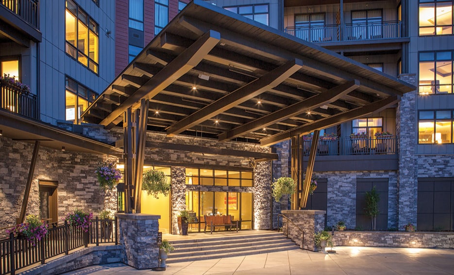 The entrance features a spacious overhang supported by wooden beams, welcoming guests with large glass doors. Stone accents and vibrant flower arrangements add to the exterior's appeal, while warm lighting enhances visibility for evening arrivals.
