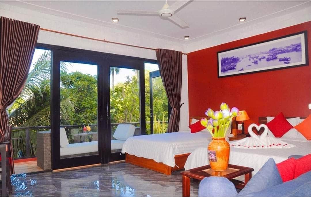 A spacious bedroom features two beds with colorful cushions and a decorative vase holding fresh flowers. Large glass doors open to a balcony, with views of lush greenery. Natural light brightens the room, highlighting the contemporary decor and polished flooring.