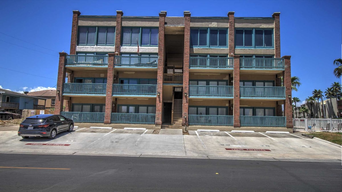 A three-story condo building is shown, constructed with a blend of brick and light-colored materials. Each level features multiple balconies with blue railings. A parking area is visible in front, with a single vehicle parked. Clear blue skies and palm trees enhance the coastal ambiance.